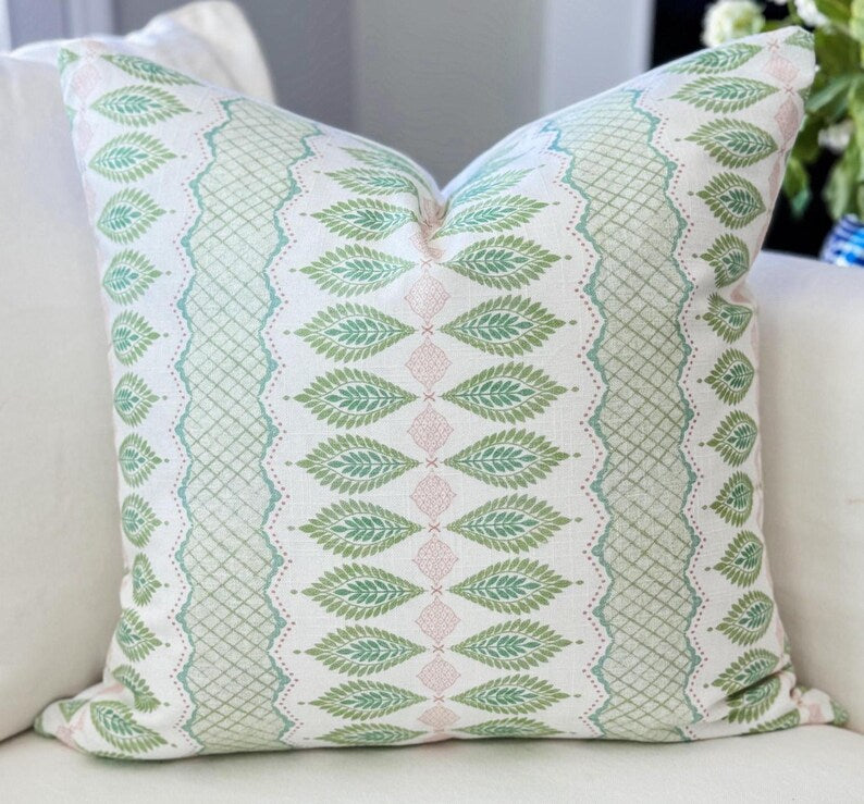 A square throw pillow with a green and pink bohemian-style block print pattern on an off-white background, resting on a light beige sofa.