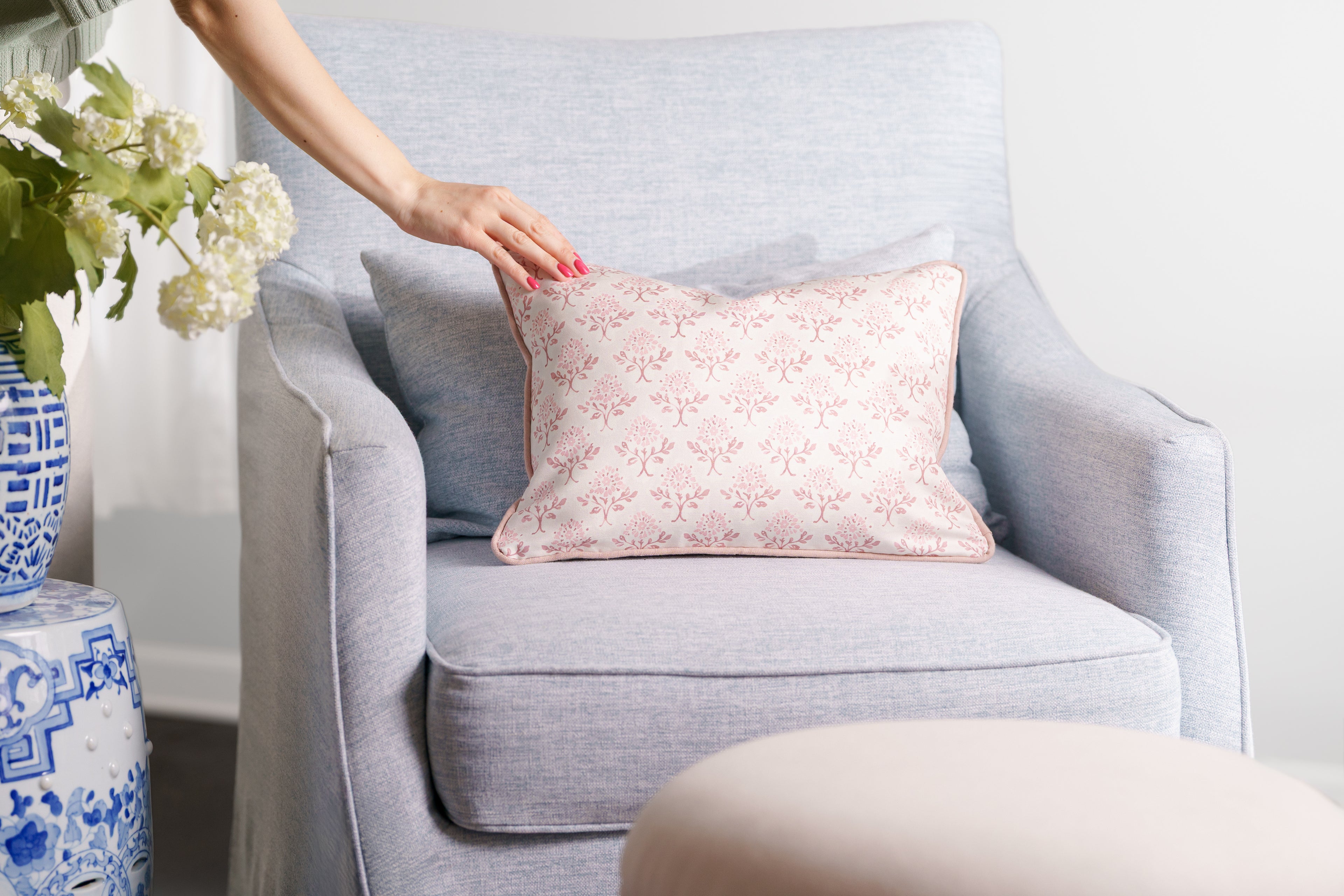 Person arranging a pink floral pillow on a gray sofa with a vase of flowers and another pillow in the background.
