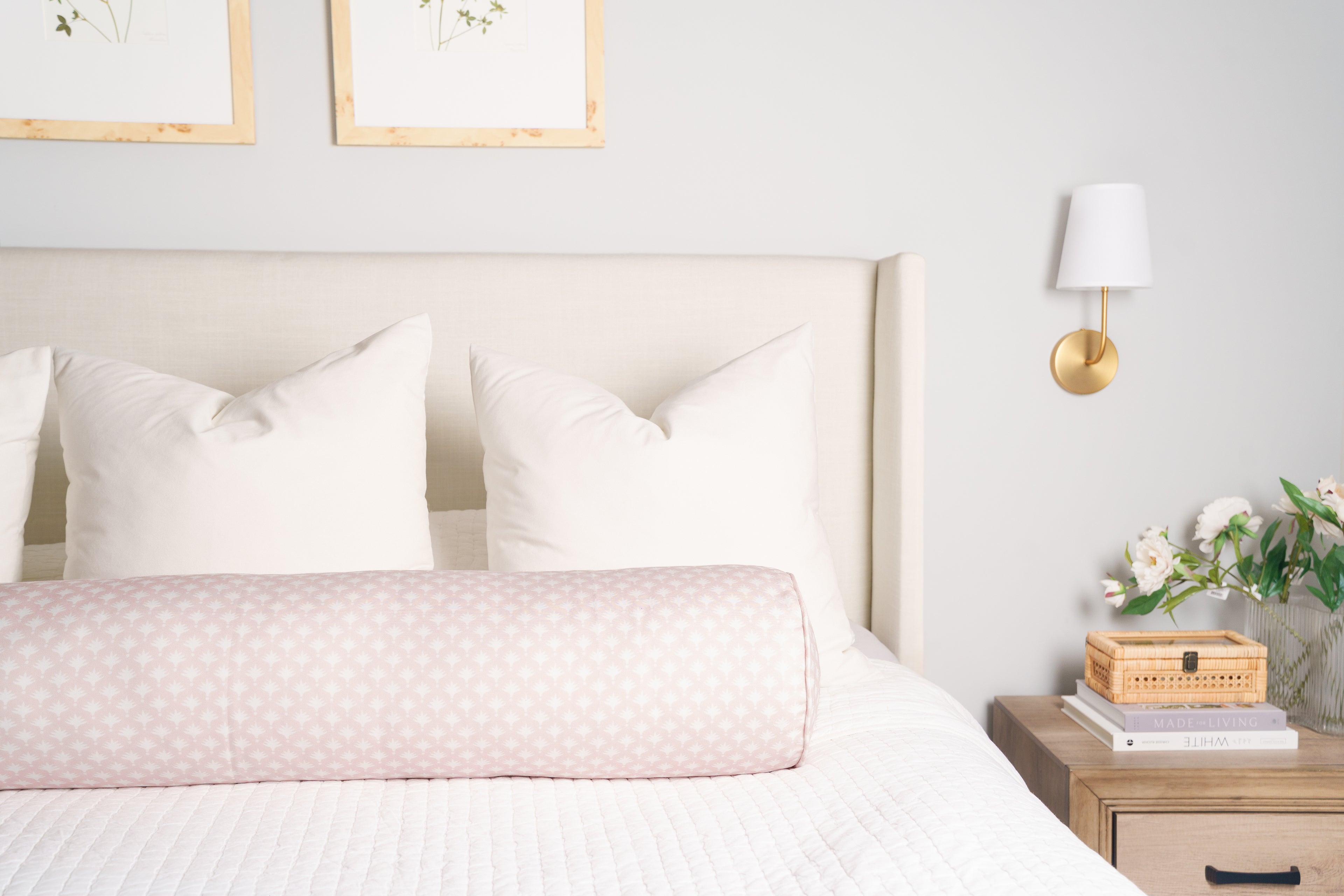 Neatly made bed with white bedding and pink decorative pillow, nightstand with books and flowers, framed pictures on wall.